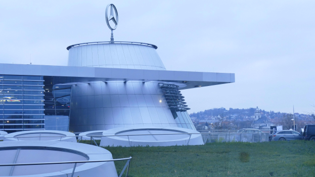 Das Mercedes-Benz Museum in Frankfurt, Deutschland, ein großes Gebäude mit seinem Logo auf dem Dach, umgeben von Gras und Geländern, mit Fahrzeugen auf der Straße davor und Bäumen im Hintergrund unter einem klaren blauen Himmel.