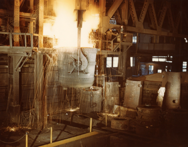 Großes Metallobjekt wird in einen Ofen in einer Fabrik gegossen, umgeben von Arbeitern, beleuchtet vom Ofenfeuer und mit Metallobjekten, Polen und Fenstern im Hintergrund.