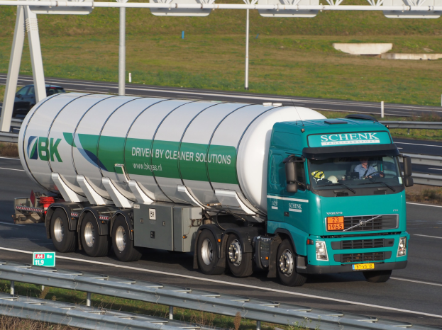 Großer Tanklastzug auf einer Autobahn mit einem sichtbaren Fahrer, umgeben von Geländern und Gras, mit Beschilderung im Hintergrund.