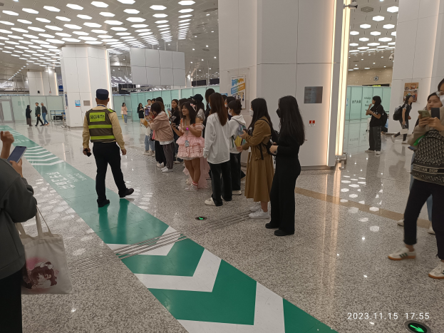 Menschen, die in einer Reihe an einem Flughafen-Sicherheitscheck-in stehen, einige mit Taschen und Mobiltelefonen, mit Säulen und Deckenleuchten im Hintergrund.