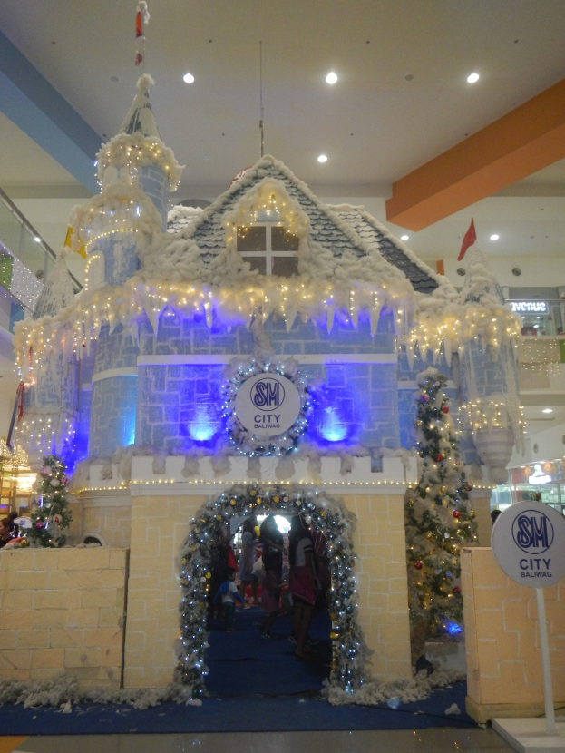 Ein festlich geschmücktes Weihnachtsdorf mit einem burgenähnlichen Aufbau, umgeben von Menschen in einem Einkaufszentrum mit beleuchteter Decke und einer Texttafel auf der rechten Seite.