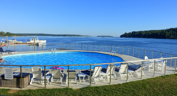 Gro├čer Schwimmbad mit Liegestühlen und Whirlpool am Grünen Ufer, umgeben von einem Gel├Ąnder, mit Booten auf einem See, B├Ąumen und einem klaren blauen Himmel im Hintergrund.