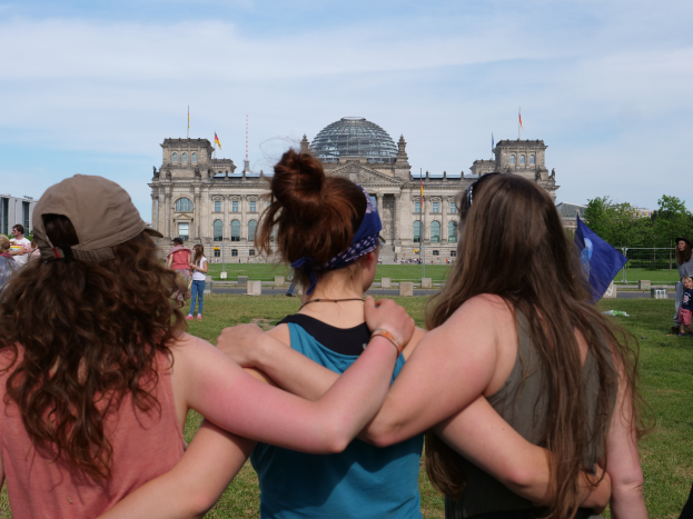 Drei Frauen in verschiedenen farbigen Kleidern, eine trägt eine Mütze, stehen vor dem Reichstaggebäude in Berlin, Deutschland, mit Menschen, Fahnen, Bäumen, Wolken und einem bewölkten Himmel im Hintergrund.