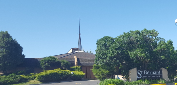 St. Bernard Catholic Church in San Antonio, Texas, ein Gebäude mit einem Kreuz darauf, umgeben von Pflanzen, Gras, Bäumen, einem Straßenpfahl, einem Schild und Straßenlaternen.