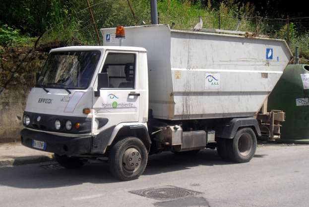 Ein weißer Müllwagen steht am Straßenrand mit einem grünen Müllcontainer rechts daneben, Bäume und eine Wand im Hintergrund.