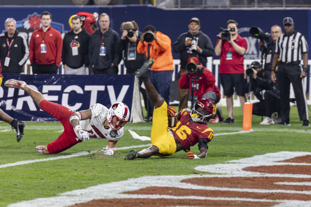 Ein Football-Spieler taucht in die Endzone ein, um einen Touchdown zu erzielen, mit Zuschauern und Medien im Hintergrund und einem Banner.