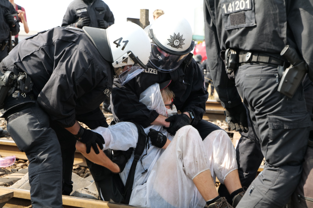 Eine Gruppe von Polizisten in Schutzausrüstung hält einen Mann in einem weißen Kleid auf einem Bahngleis fest, mit anderen Personen im Hintergrund.