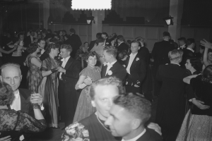 Eine Gruppe von Menschen, die auf einer Party in einem Schwarz-Wei├č-Setting tanzen, mit M├Ąnnern und Frauen verschiedenen Alters, einige halten Gl├Ąser, und eine beleuchtete Wand im Hintergrund.