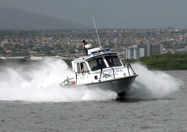 Polizeiboot fährt durch Wasser in der Nähe einer Stadt, mit sichtbaren Insassen, vor dem Hintergrund von Bäumen, Gebäuden, Strommasten, Bergen und einem klaren Himmel.