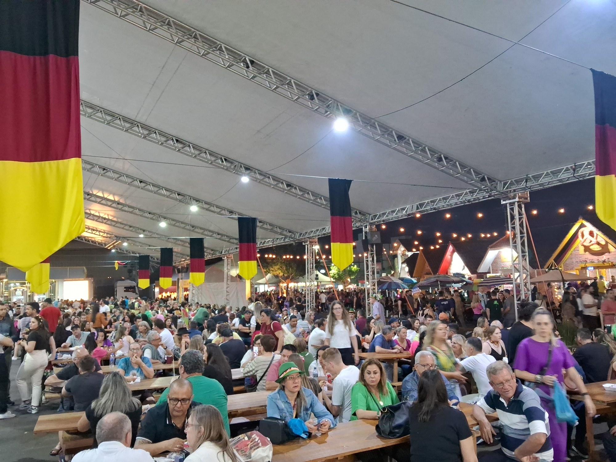 Eine große Gruppe von Menschen sitzt an Tischen unter einem Zelt auf dem Oktoberfest in München, Deutschland, einige stehen und Taschen liegen auf den Tischen, vor einem festlichen Hintergrund aus Zelten, Lichtern und Fahnen.