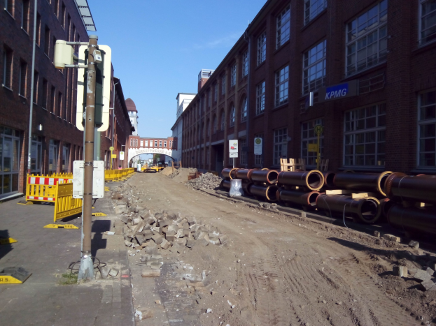 Großer Haufen von Rohren an einer Baustelle neben einer Straße mit Gebäuden, Schildern, Metallpfosten, Absperrungen, Steinen und einem Bogen im Hintergrund unter einem bewölkten Himmel.