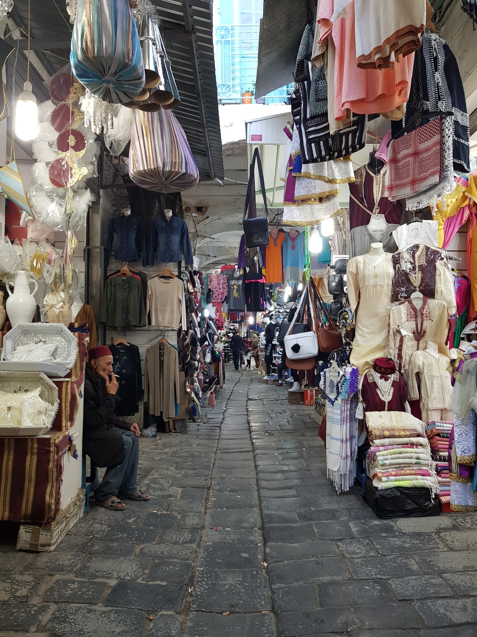 Ein belebter Straßenmarkt in Jerusalem mit Ständen, die Kleidung, Taschen, Lampen und andere Artikel verkaufen, umgeben von Menschen, die durch die Stände schlendern und ein Gebäude im Hintergrund.