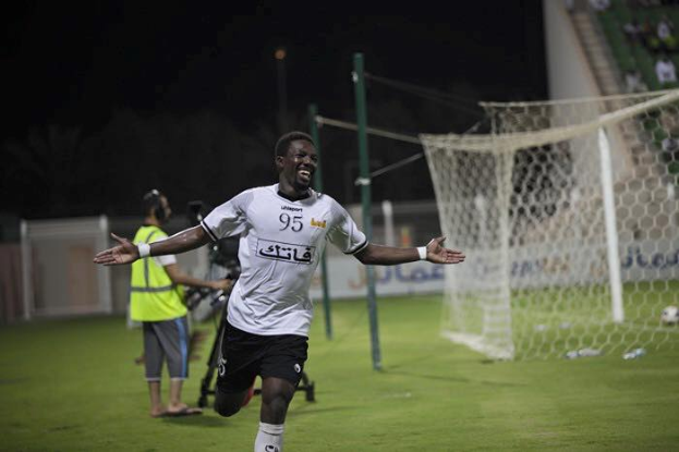 Mann in weißem Hemd und schwarzen Shorts rennt auf einem Fußballfeld mit ausgestreckten Armen, feiernd, umgeben von Menschen, mit einem Torpfosten rechts und Zuschauern im Hintergrund.