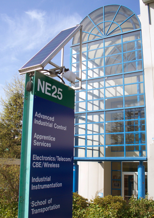 Gebäude mit Glasfenstern und -tür, Außenschilder, Dachsolarpanel, umliegende Pflanzen und Bäume, bewölkter Himmel.