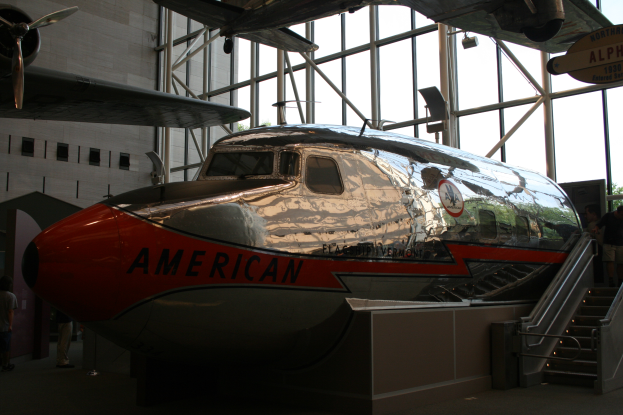 Ein American-Airlines-Flugzeug in einem Museum mit Menschen drumherum, Treppe auf der rechten Seite und einem Gebäude, Bäumen und einem klaren blauen Himmel im Hintergrund.