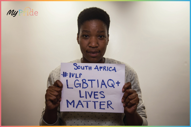 Frau in weißem Hemd mit einem Schild, auf dem 'LGBTQ+ Lives Matter' steht, vor einer Wand.