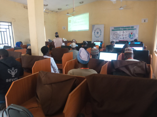 Eine Gruppe von Menschen, die an Computern in einem Seminarrraum mit Deckenventilatoren, einem Bildschirm, einem Banner und Fenstern auf beiden Seiten sitzen.