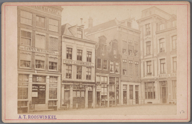 Ein altes Foto einer Stadtstraße mit Gebäuden, die Fenster, Türen und Namensschilder aufweisen, mit Text am unteren Bildrand.