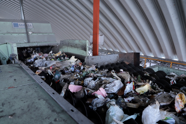 Großer müllhaufen auf einem förderband in einer recycling-anlage mit maschinentechnik und einem metallenen dach darüber.