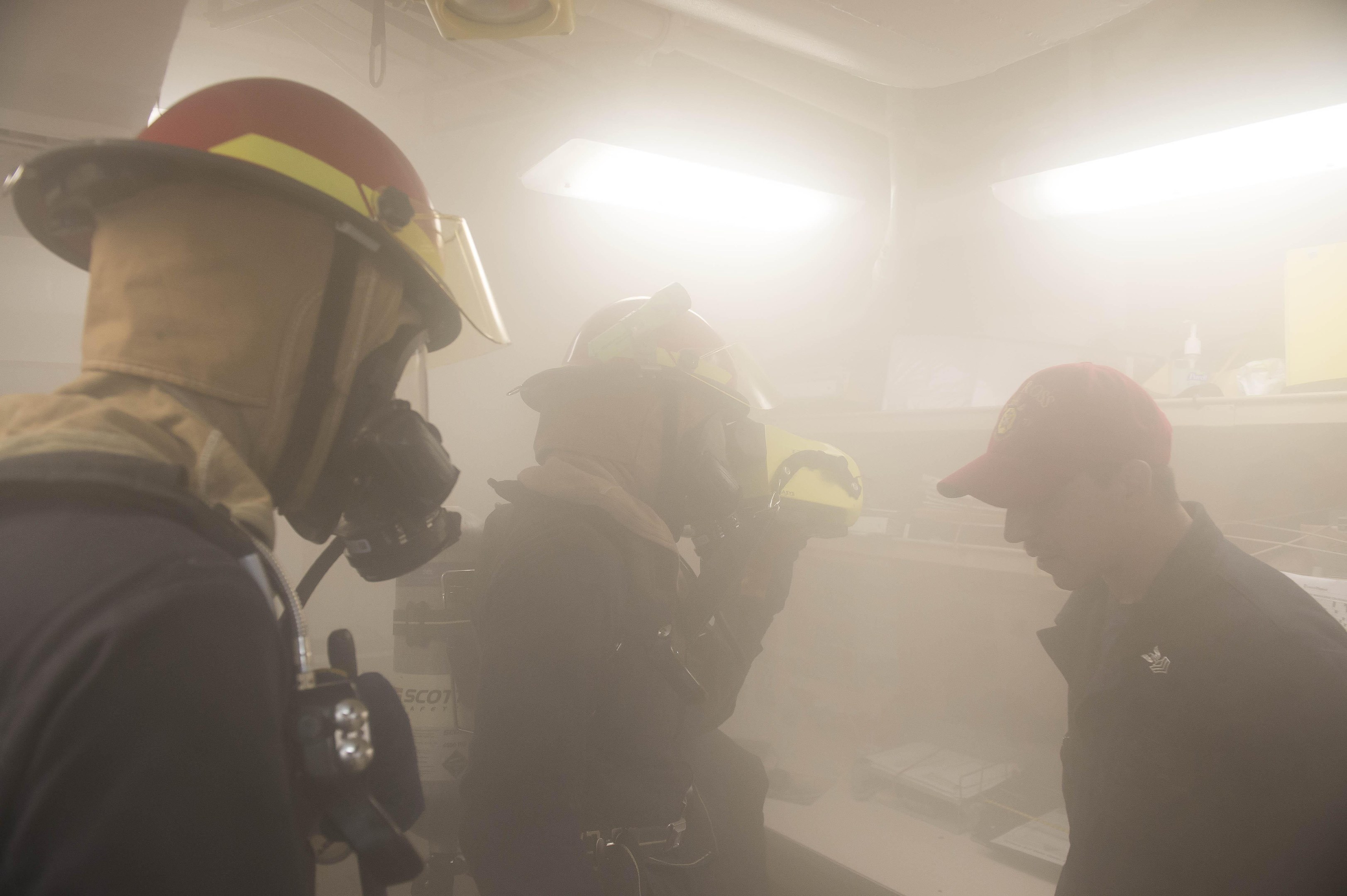 Feuerwehrleute in Helmen mit Rauch aus dem Mund, einer hält eine Kamera, während einer Trainingsrunde mit Hintergrundbeleuchtung und Gegenständen.
