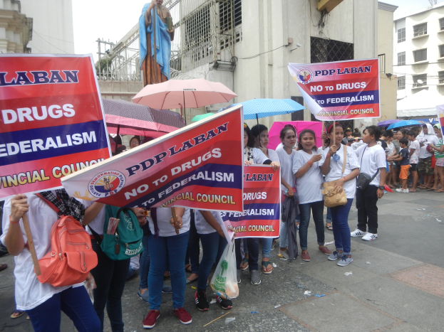 Gruppe von Menschen demonstriert vor einem Gebäude mit einer Statue im Hintergrund, hält Schilder und Schirme unter einem klaren blauen Himmel.