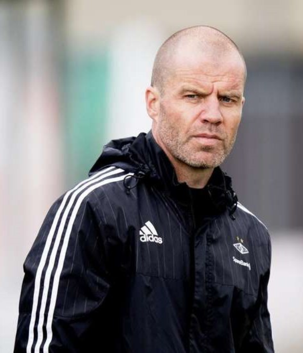 Mann in marineblauem Jackett mit weißen Streifen und weißem Kragen, der auf einem Fußballfeld steht und die Hände verschränkt hält.