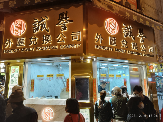 Gruppe von Menschen vor einem hell erleuchteten Gebäude nachts mit Schildern und einige mit Taschen, in der Nähe einer Geldwechsel-Geschäft.