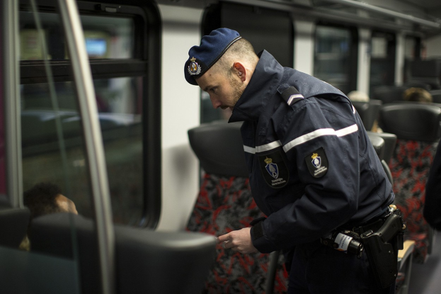 Polizeibeamter in Uniform und Mütze auf einem Zug stehend, der auf sein Handy schaut, mit sitzenden Passagieren und einer weiteren Person rechts.