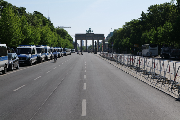 Lange Reihe von Polizeiwagen, die vor dem Brandenburger Tor auf der Straße geparkt sind, mit Fahrradfahrern und Fußgängern, Barrieren, Bäumen und einem Bogen mit Statuen im Hintergrund.