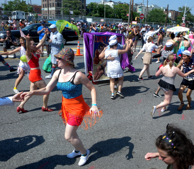 Eine Gruppe von Menschen, die eine Straße entlangmarschieren, einige mit Mötzen und Schirmen, andere tanzend, mit einem Banner, Verkehrskegeln, Fahrzeugen, Laternen, Schildern, Bäumen, Gebäuden und einem klaren blauen Himmel im Hintergrund.