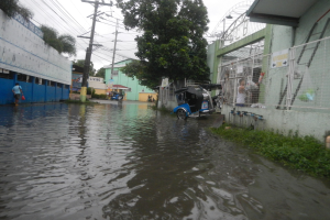 Eine überflutete Straße mit einem Motorrad in der Mitte, umgeben von Gebäuden, Bäumen und Strommasten mit Drähten, während im Hintergrund Menschen und Fahrzeuge zu sehen sind.