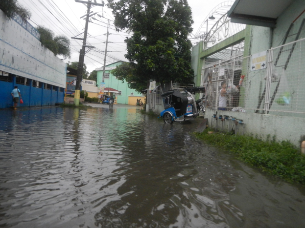 Eine überflutete Straße mit einem Motorrad in der Mitte, umgeben von Gebäuden, Bäumen und Strommasten mit Drähten, während im Hintergrund Menschen und Fahrzeuge zu sehen sind.