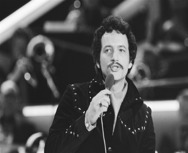 Schwarzes und weißes Foto von Elvis Presley, der ein Mikrofon während einer Live-Aufführung hält, mit einem unscharfen Hintergrund.