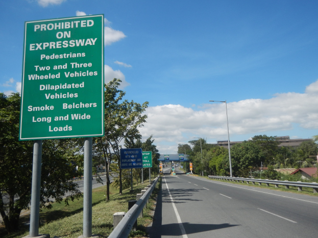 Grüne Autobahnschilder mit der Aufschrift "Verboten auf der Autobahn" mit Fahrzeugen, Laternenpfählen, Bäumen, Gebäuden und einem bewölkten Himmel im Hintergrund.