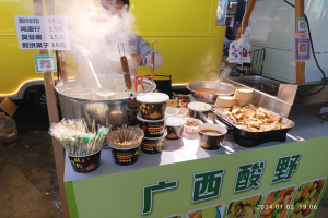 Ein Mann steht vor einem chinesischen Imbisswagen und bereitet Gerichte mit Schalen, Tassen, Löffeln, Zangen und anderen Utensilien auf der Theke vor.