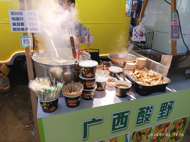 Ein Mann steht vor einem chinesischen Imbisswagen und bereitet Gerichte mit Schalen, Tassen, Löffeln, Zangen und anderen Utensilien auf der Theke vor.