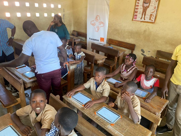 Kinder sitzen an Tischen und nutzen Tablets in einem Klassenzimmer mit Erwachsenen in der Nähe, ein Banner und ein Plakat an der Wand.