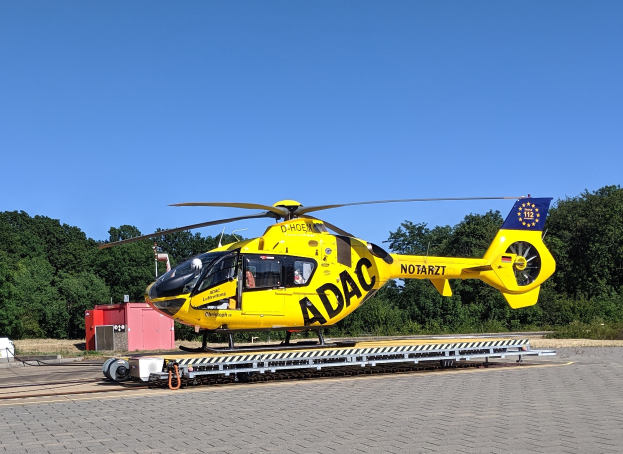 Gelber Hubschrauber auf einem Flughafenvorfeld mit Bäumen und einem klaren blauen Himmel, wahrscheinlich ein Luftrettungshubschrauber in Deutschland.