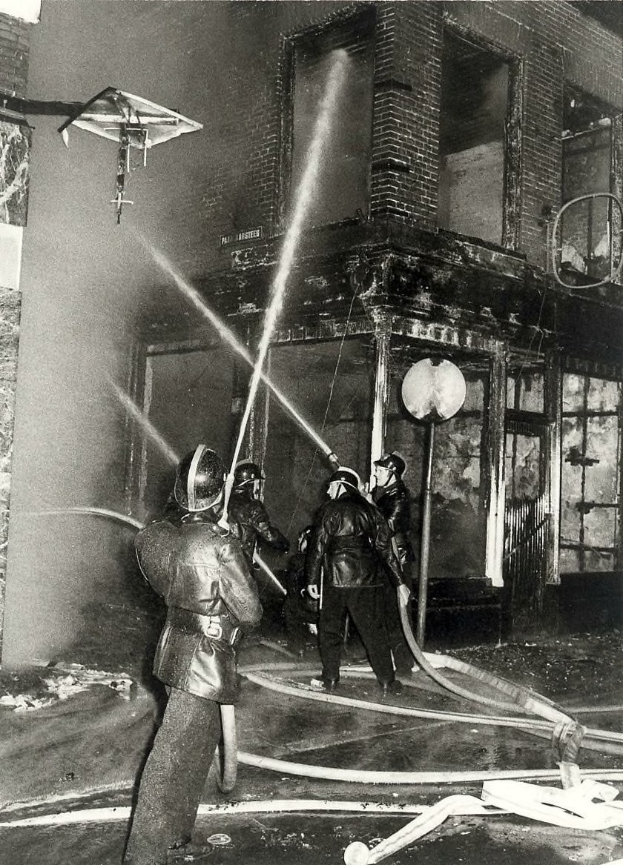 Schwarzes und weißes Foto von Feuerwehrleuten in Helmen mit Schläuchen in den Händen, die vor einem feuerbeschädigten Gebäude stehen.