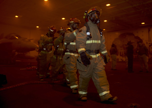 Feuerwehrleute in Schutzausrüstung durchqueren einen Hangar mit einem Flugzeug und Deckenbeleuchtung im Hintergrund, während auf dem Boden verstreute Gegenstände zu sehen sind.