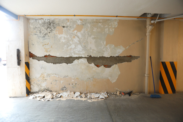 Leere Tiefgarage mit abblätternder Farbe an den Wänden, verstreute Papiere und Schutt auf dem Boden, ein Besenstiel, ein Pfahl und Rohre an der Decke, mit einem Fahrzeug auf der linken Seite sichtbar.