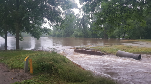 Überschwemmte Straße mit einem großen Rohr in der Mitte, umgeben von Gras und Bäumen, unter einem sichtbaren Himmel, mit einem Wasserzeichen.