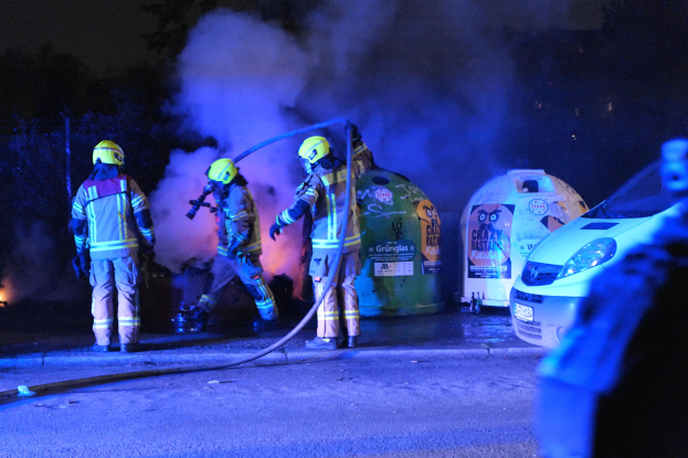 Feuerwehrleute mit Helmen auf einer Straße mit einem brennenden Auto, Rauch aufsteigend, Bäume und ein Zaun im Hintergrund.