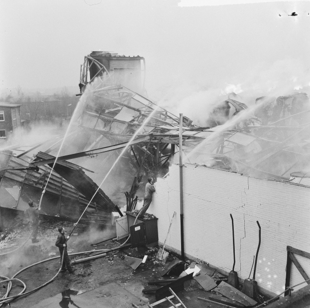 Schwarzes und weißes Foto von Feuerwehrleuten auf dem Dach eines Gebäudes, die ein Feuer löschen, während Menschen unten mit Ausrüstung helfen.