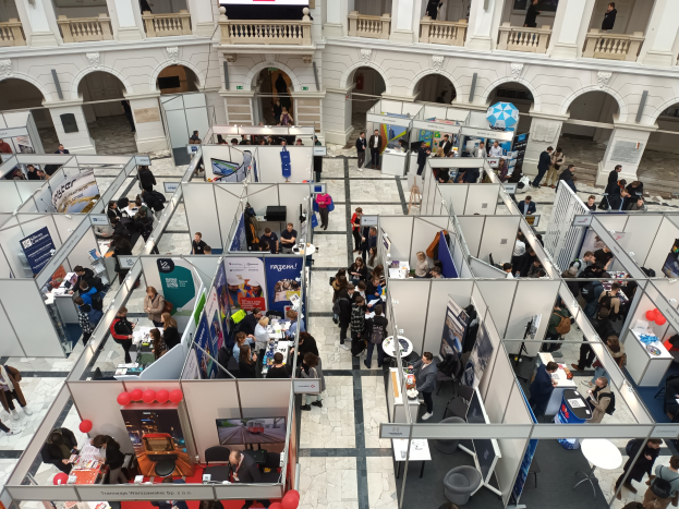 Große Besuchergruppe auf einer internationalen Fachmesse mit Ständen, Bannern, Tischen, Stühlen und Luftballons in einem Raum mit architektonischen Bögen, Säulen und einem Bildschirm im Hintergrund.