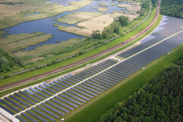 Luftaufnahme eines Solarparks mit Solarpanelen, umgeben von Bäumen, Gras, Pflanzen und Wasser, mit einem Zug auf einer Eisenbahnschiene.