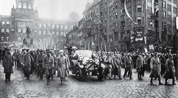 Eine Gruppe von Menschen in Uniformen, einige halten Fahnen, gehen eine Straße in Berlin während des Ersten Weltkriegs entlang, mit einem Auto in der Mitte und Gebäuden auf beiden Seiten.