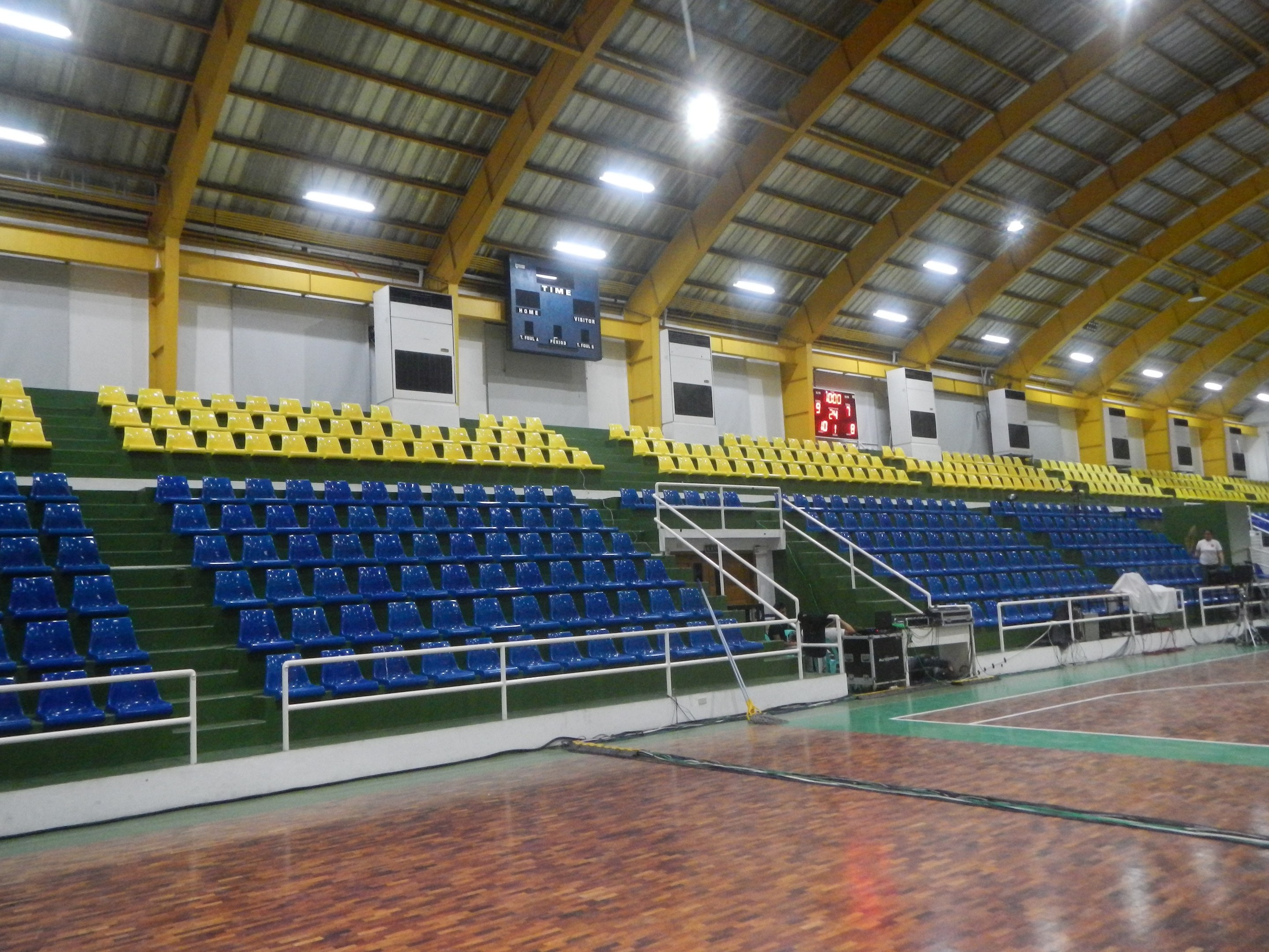 Große Sporthalle mit Holzboden, blauen und gelben Sitzplätzen, Geländern, Treppen zum Sitzbereich, Wandbildschirmen, Deckenleuchten und einigen Anwesenden.