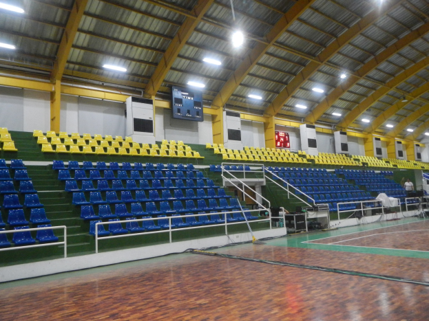 Große Sporthalle mit Holzboden, blauen und gelben Sitzplätzen, Geländern, Treppen zum Sitzbereich, Wandbildschirmen, Deckenleuchten und einigen Anwesenden.