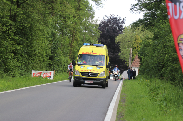 Ambulanz fährt auf einer Straße mit Radfahrern an der Seite, Gras und Bäume auf beiden Seiten, Häuser und Masten im Hintergrund bei einem klaren blauen Himmel.